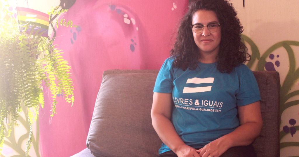 Foto da Ananda sentada num sofá encostado na parede. Ela está com camiseta da campanha Livres e Iguais da ONU.